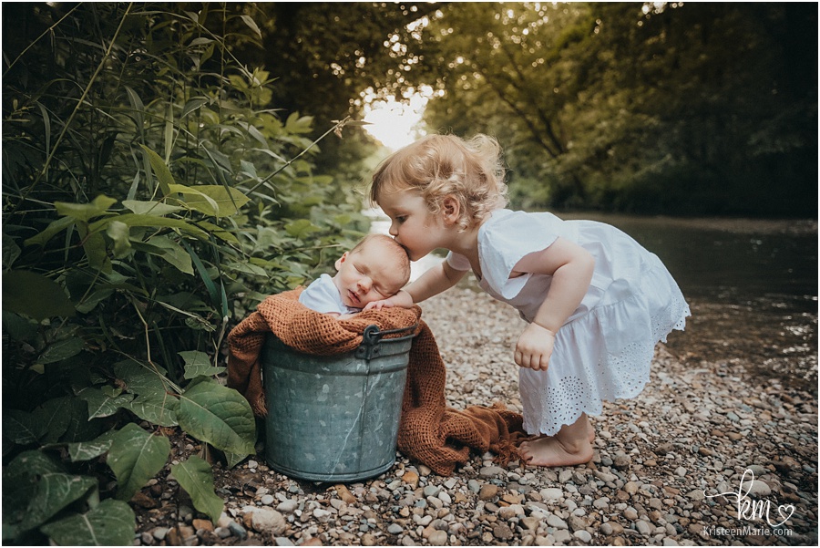 outdoor newborn picture with siblins - baby in a bucket with big sister kissing