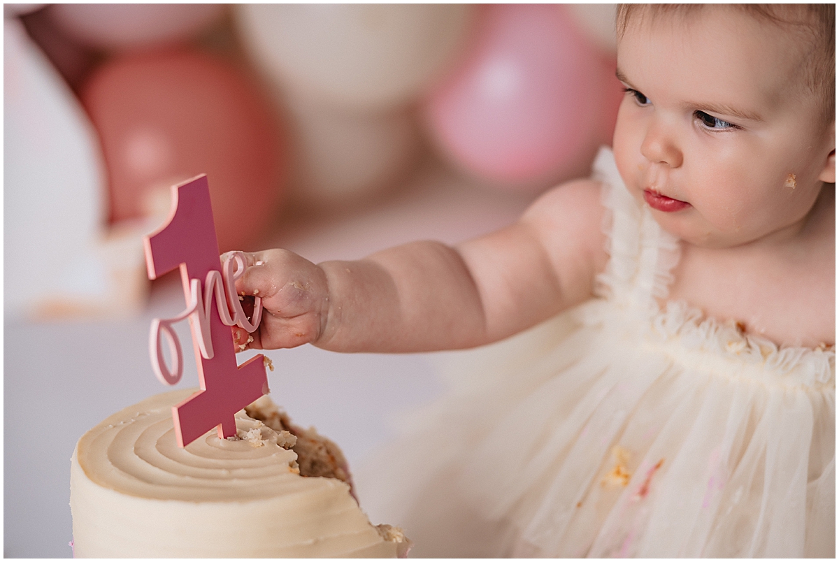 Zionsville cake smash photographer baby girl standing next to smashed cake with balloon backdrop