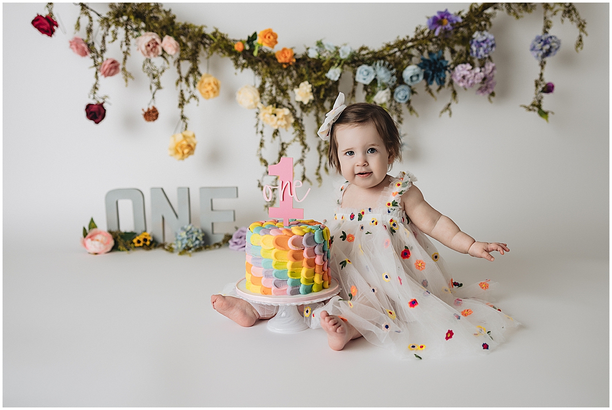 Rainbow baby sitting beside a pastel rainbow cake and floral first birthday backdrop
