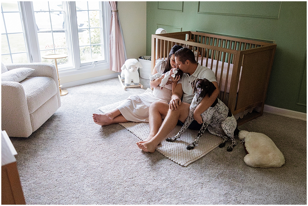 Parents sit with their newborn and dog on the nursery floor.