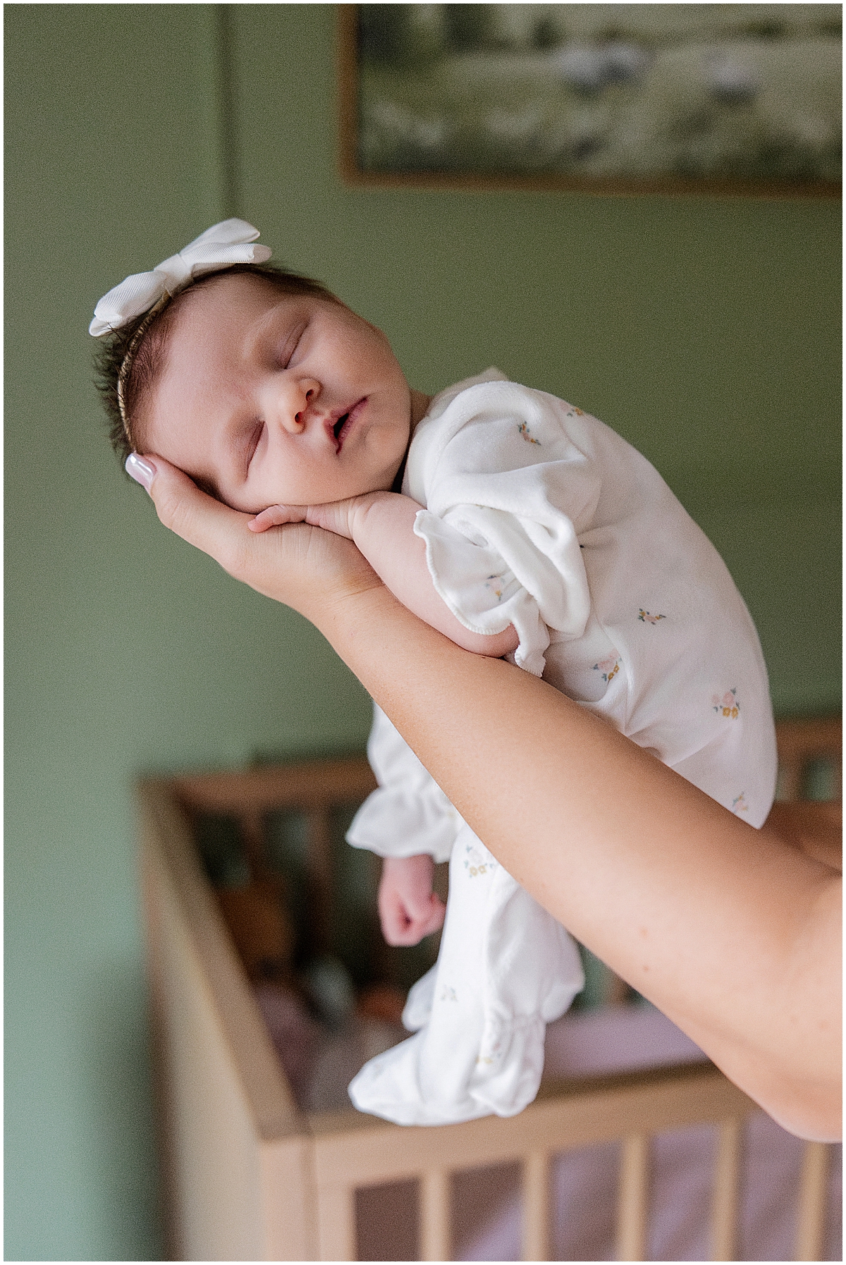 Sleeping newborn girl in a white outfit held above her nursery crib.