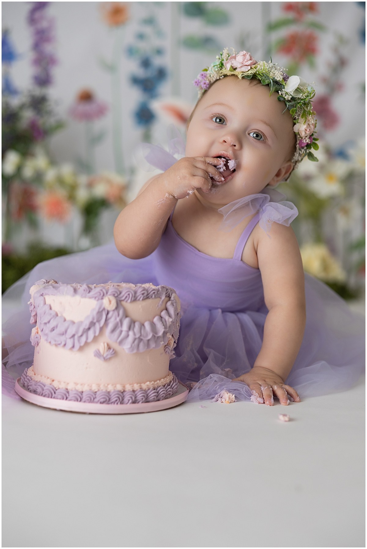 Baby tasting frosting from her fingers beside a pink cake with purple decorations.