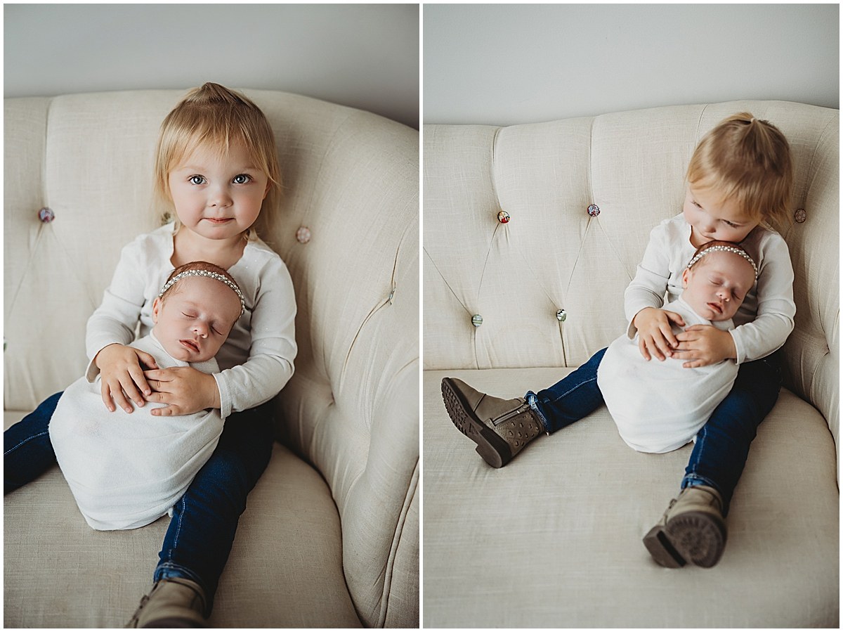 Older sister holding and kissing her wrapped newborn sister on a cream couch.