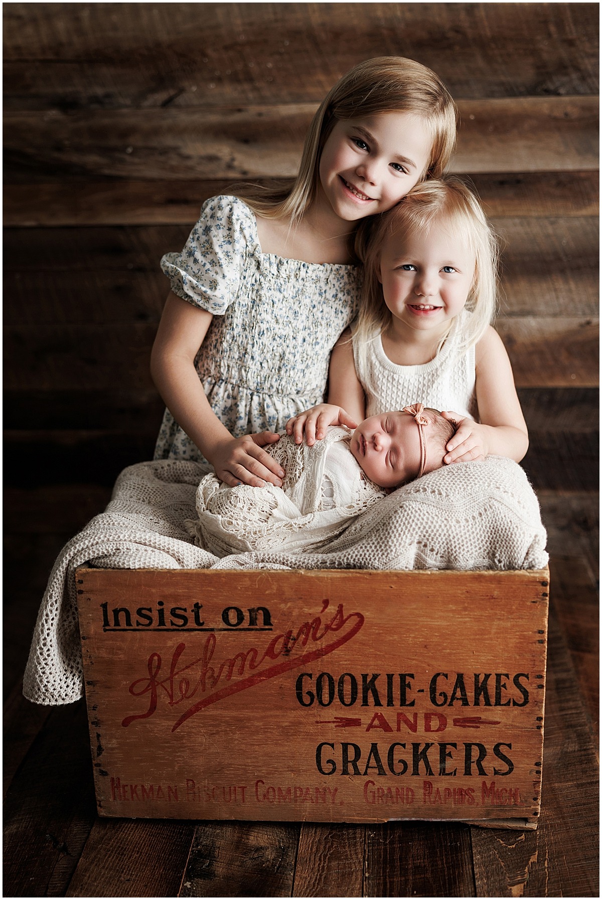 Two older sisters pose beside their newborn sister inside a wooden heirloom box bearing their family name.