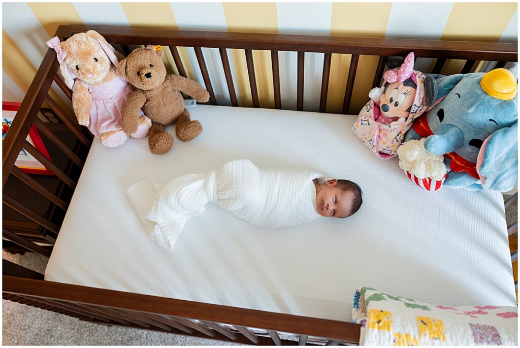 Newborn baby wrapped in white lying in a wooden nursery crib with stuffed animals.