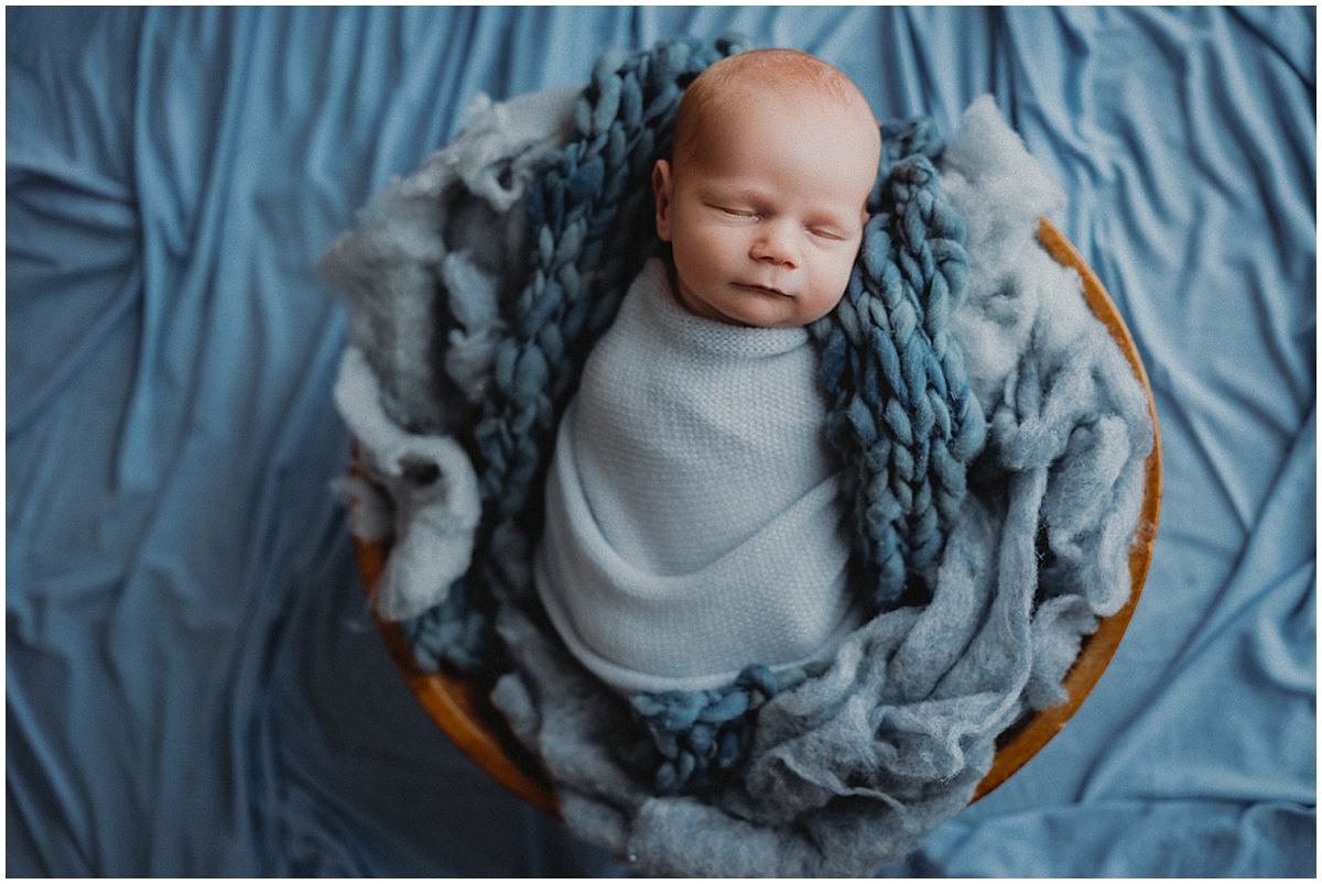 Sleeping newborn wrapped in pale blue in a wooden bowl lined with chunky blue knit fabric.