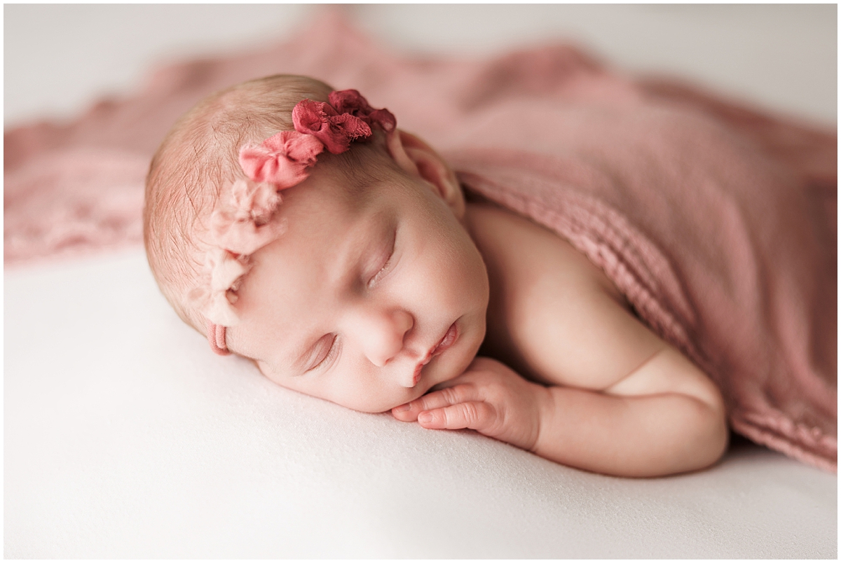Sleeping newborn resting on folded hands with a blush wrap and pink floral headband.