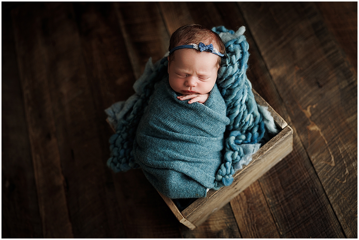 Sleeping newborn girl wrapped in blue and posed inside a rustic wooden box.