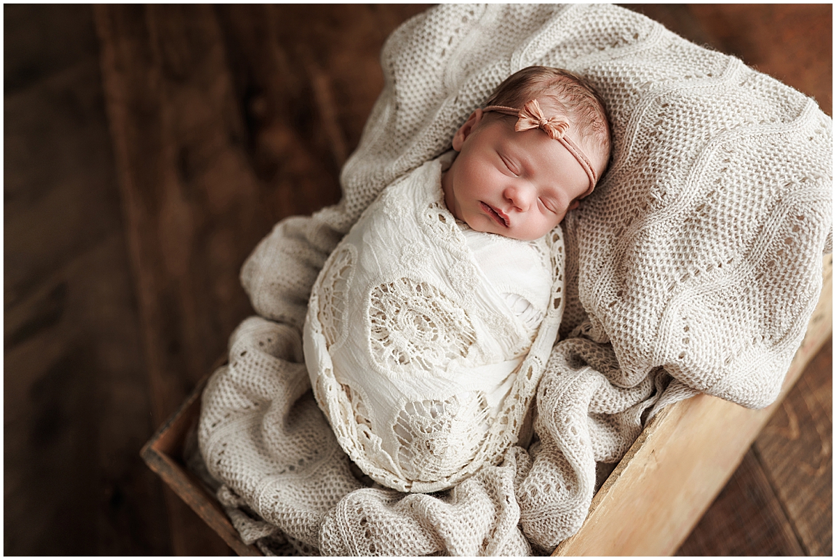 Sleeping newborn girl wearing a cream lace wrap and pink headband inside a wooden box.