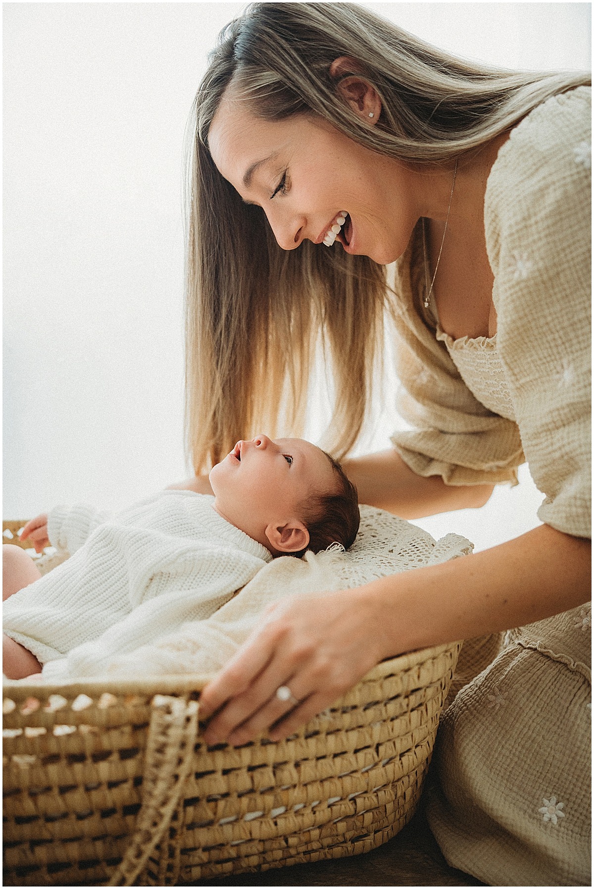 other smiling down at her newborn resting in a woven basket beside a bright window.