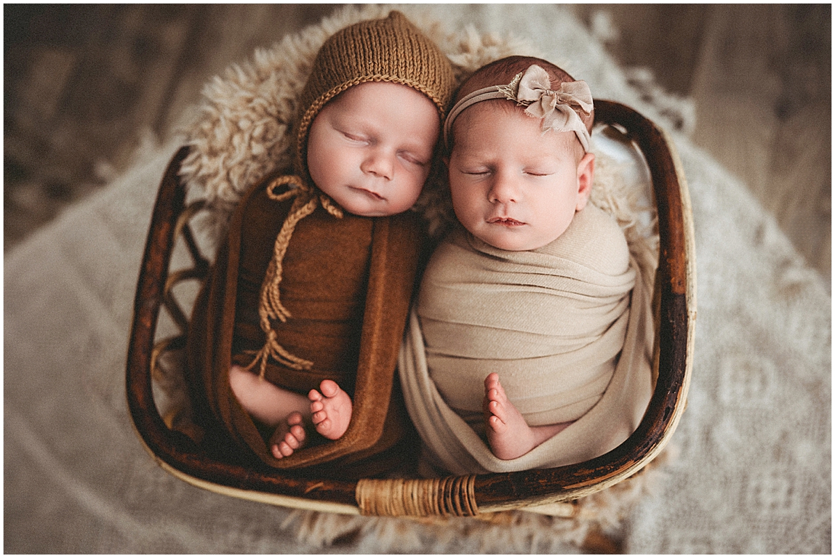 Newborn twins in brown and cream wraps resting in a rustic basket with their tiny feet visible.