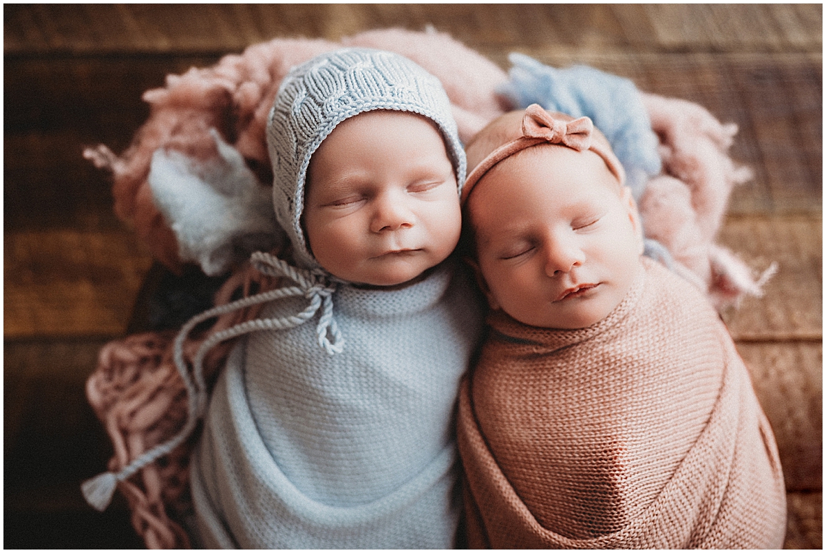 Newborn twins lying side by side in pale blue and blush wraps, with a knit bonnet and bow headband.