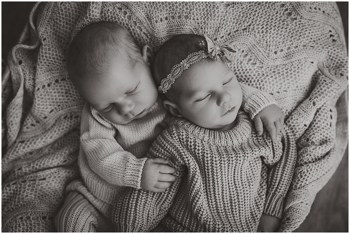 Black-and-white portrait of twins nestled together in knit outfits, with one baby’s arm around the other.