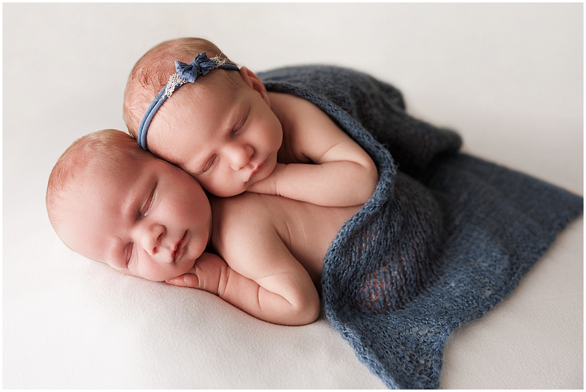 Sleeping twins nestled together beneath a navy knit blanket on a cream backdrop.