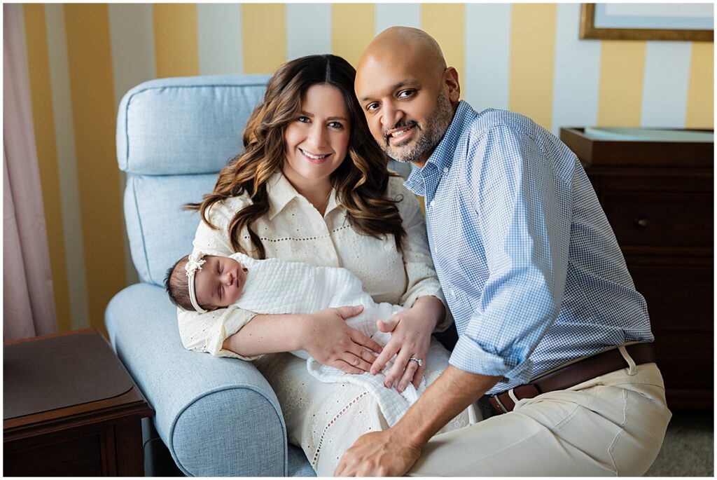Parents holding their sleeping newborn baby together on a blue nursery chair.