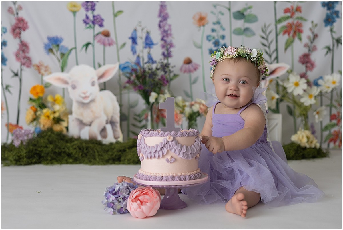 Smiling birthday girl in a purple dress beside a pink and purple cake with a floral and lamb backdrop.