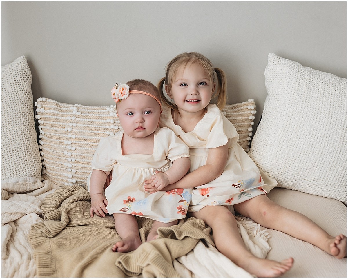 Older sister hugging her baby sister as they sit together in coordinating floral outfits.