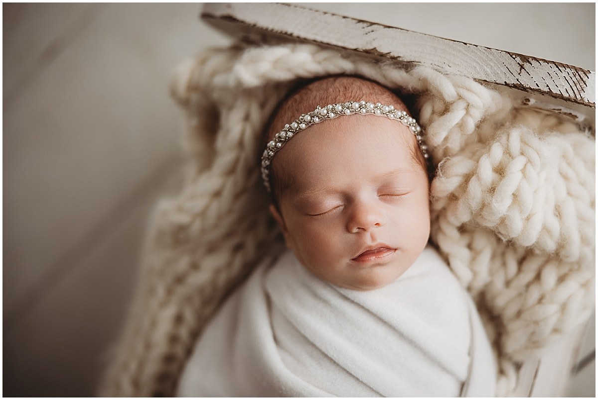 Sleeping newborn wearing a delicate headband and white wrap on a cream knit blanket.