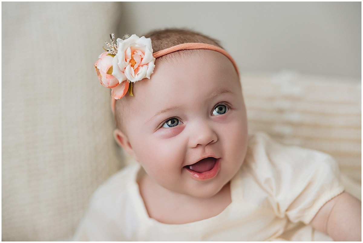 Close-up of a smiling baby wearing a peach flower headband and cream outfit.