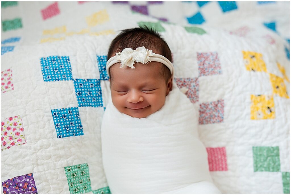 Smiling newborn baby wrapped in white on a colorful patchwork quilt.
