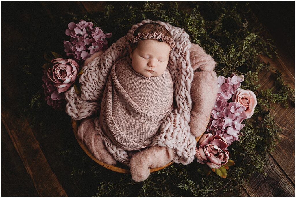 Sleeping newborn baby in a pink wrap surrounded by mauve flowers and greenery.