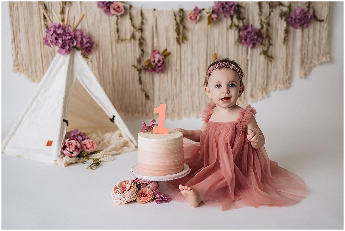 Baby girl in a pink dress beside a first birthday cake in a floral cake smash setup