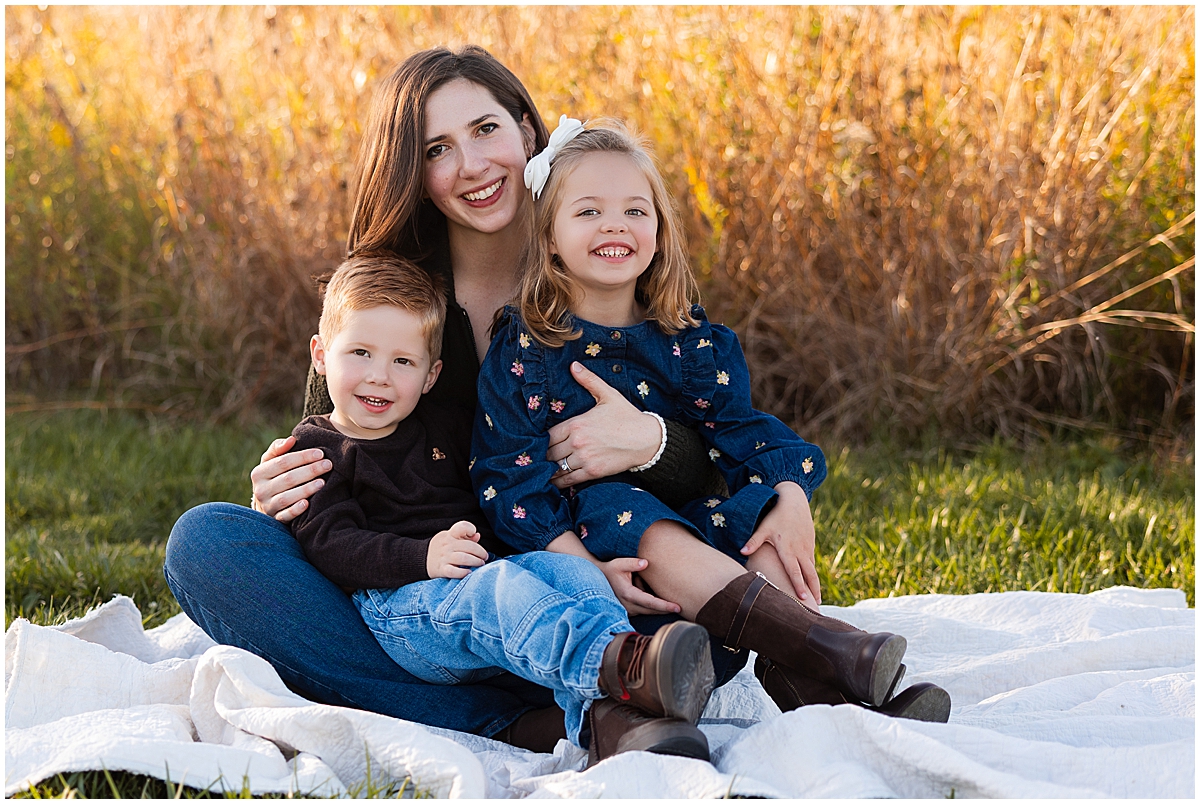 Mother cuddles her two young children on a white blanket in warm evening light.
