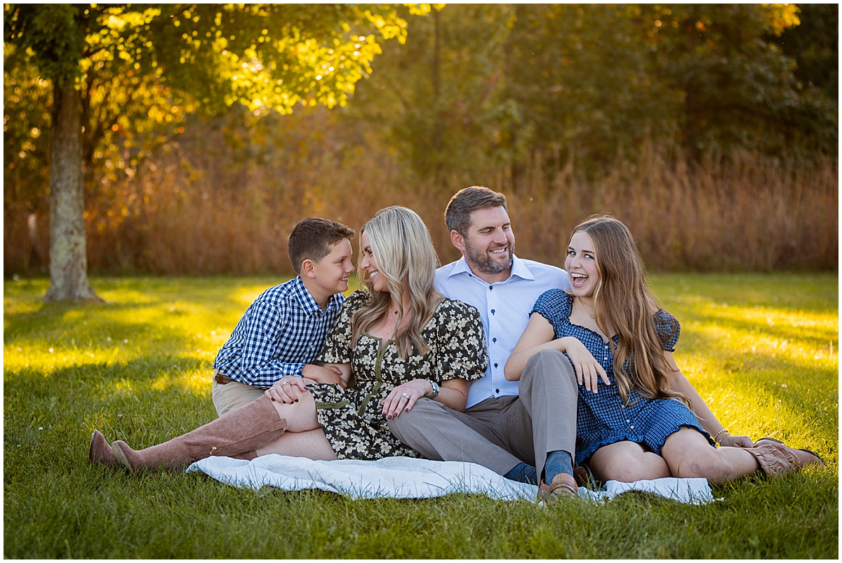 Family of four laughs together while sitting on a blanket beneath autumn trees.