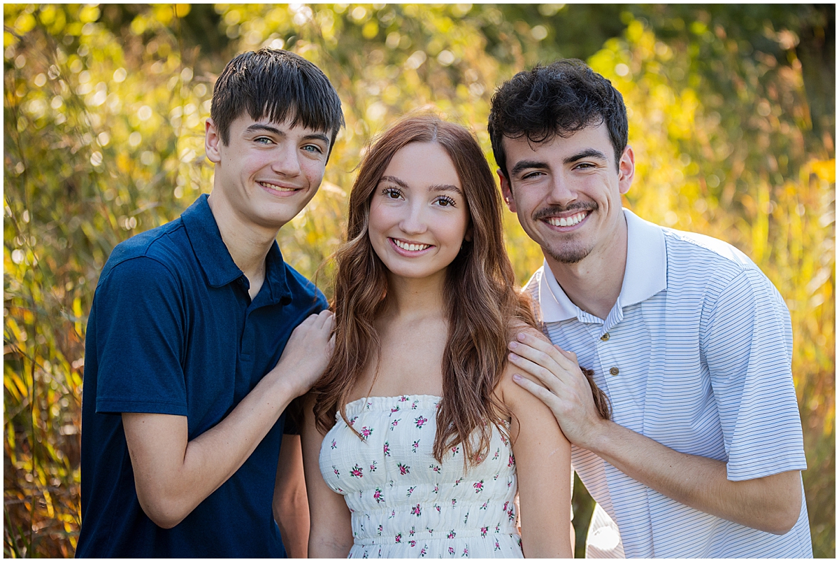 Three older siblings smile together in front of sunlit grasses.