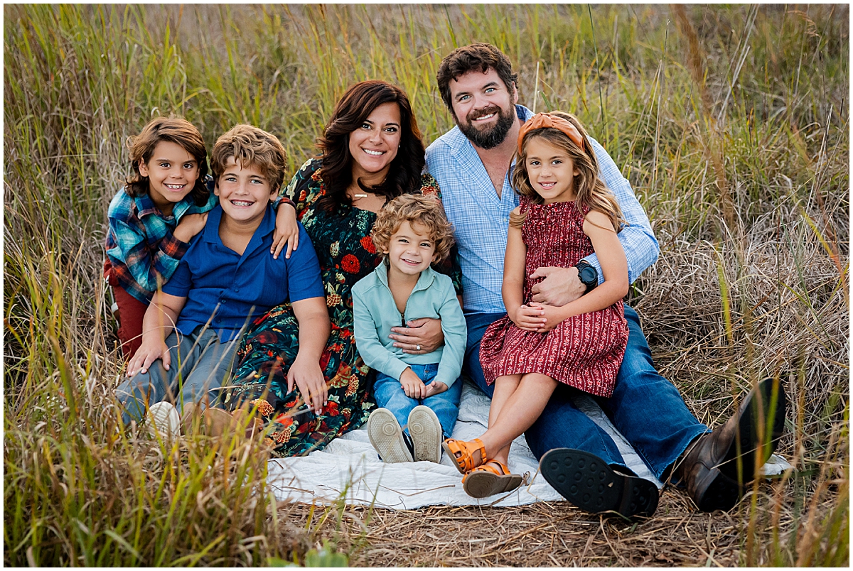 Parents and their four children sit together on a blanket among tall grasses.