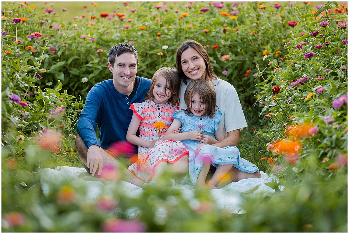 Family of four cuddling on a white blanket surrounded by colorful flowers at Dull’s Tree Farm.