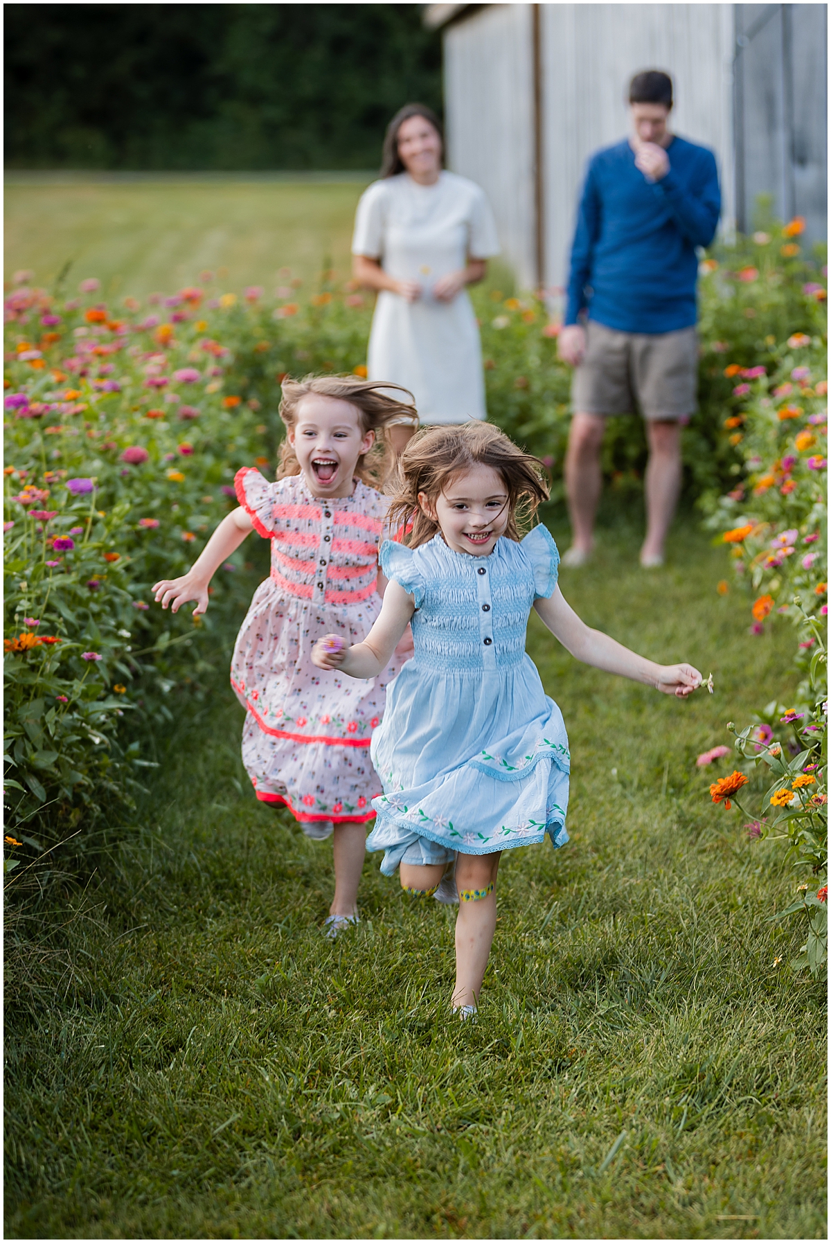 Two girls in blue and floral dresses running along a grassy path between flowers.