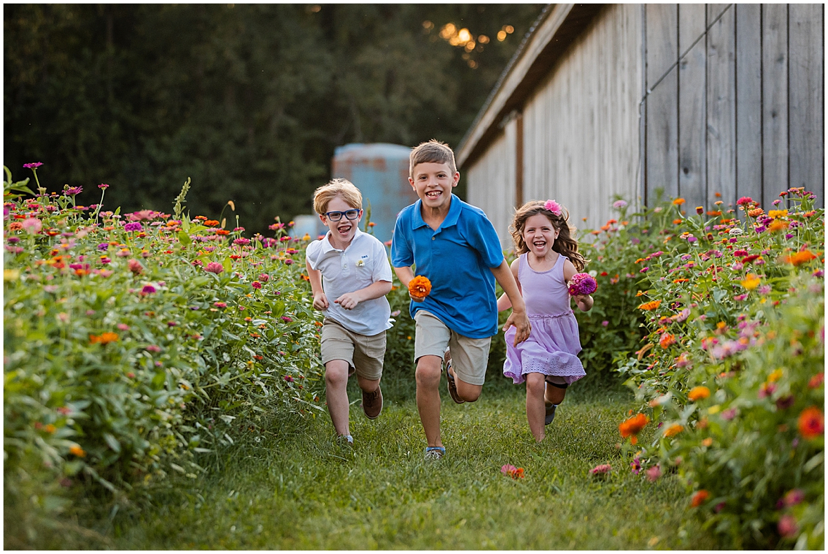 Three smiling children running toward the camera along the flower-lined path beside the barn.