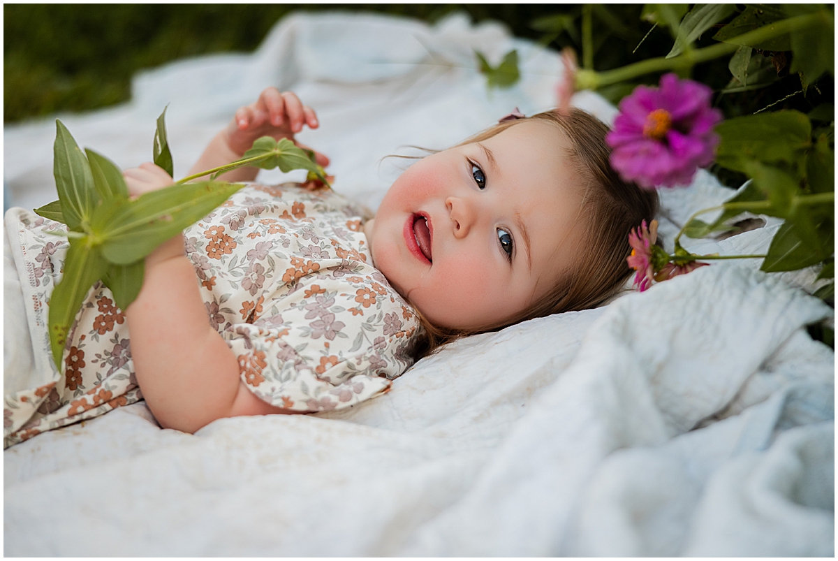 Toddler lying on a white blanket beside purple flowers and holding a flower stem.