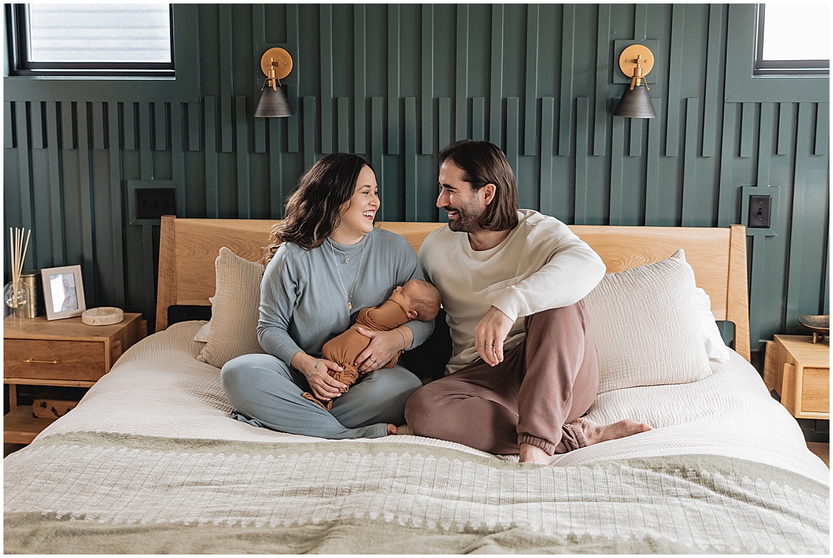 Parents smiling at each other while holding their newborn on cream bedding in a green bedroom