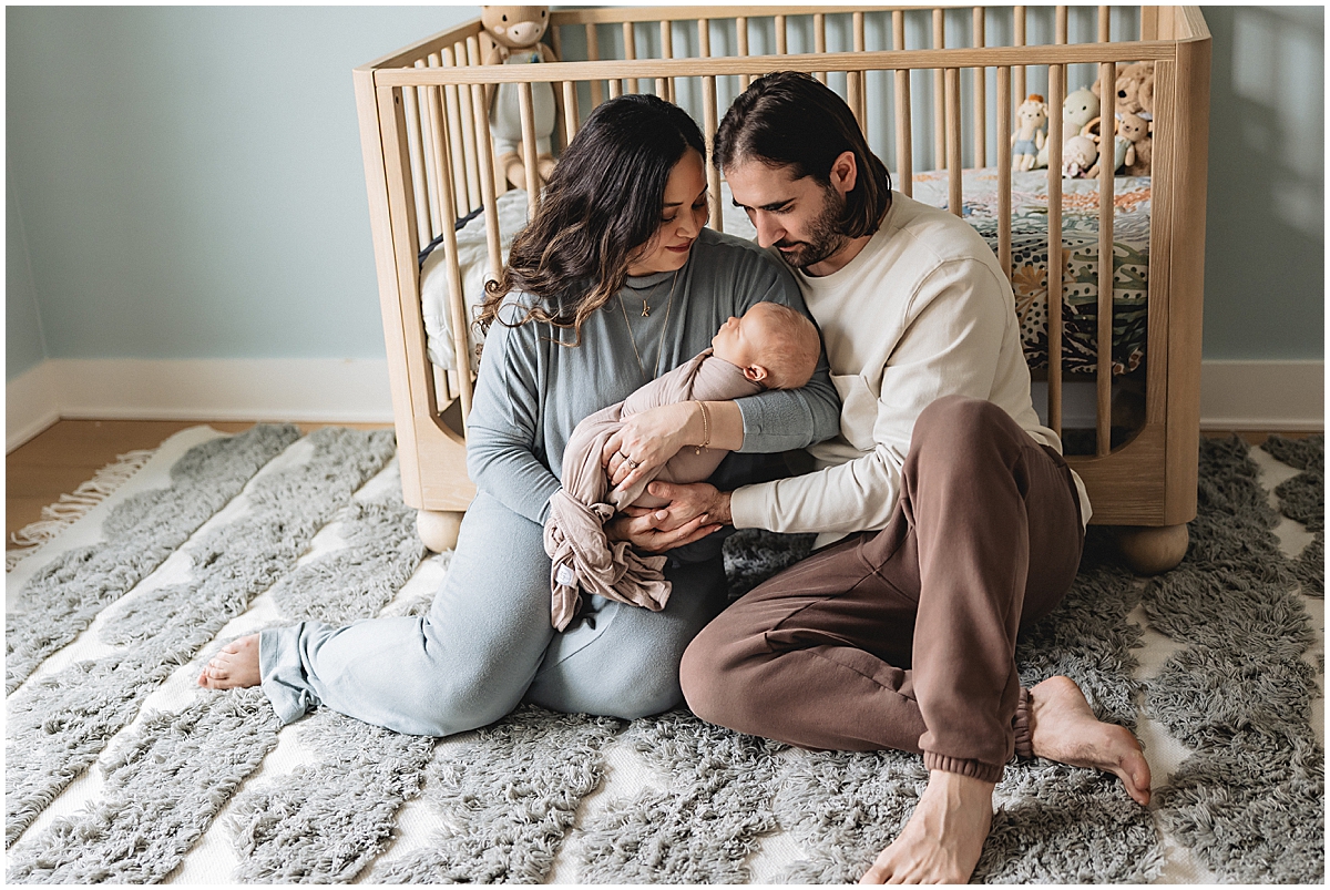Parents in coordinated blue, cream, and brown outfits sitting with their newborn beside a wooden crib