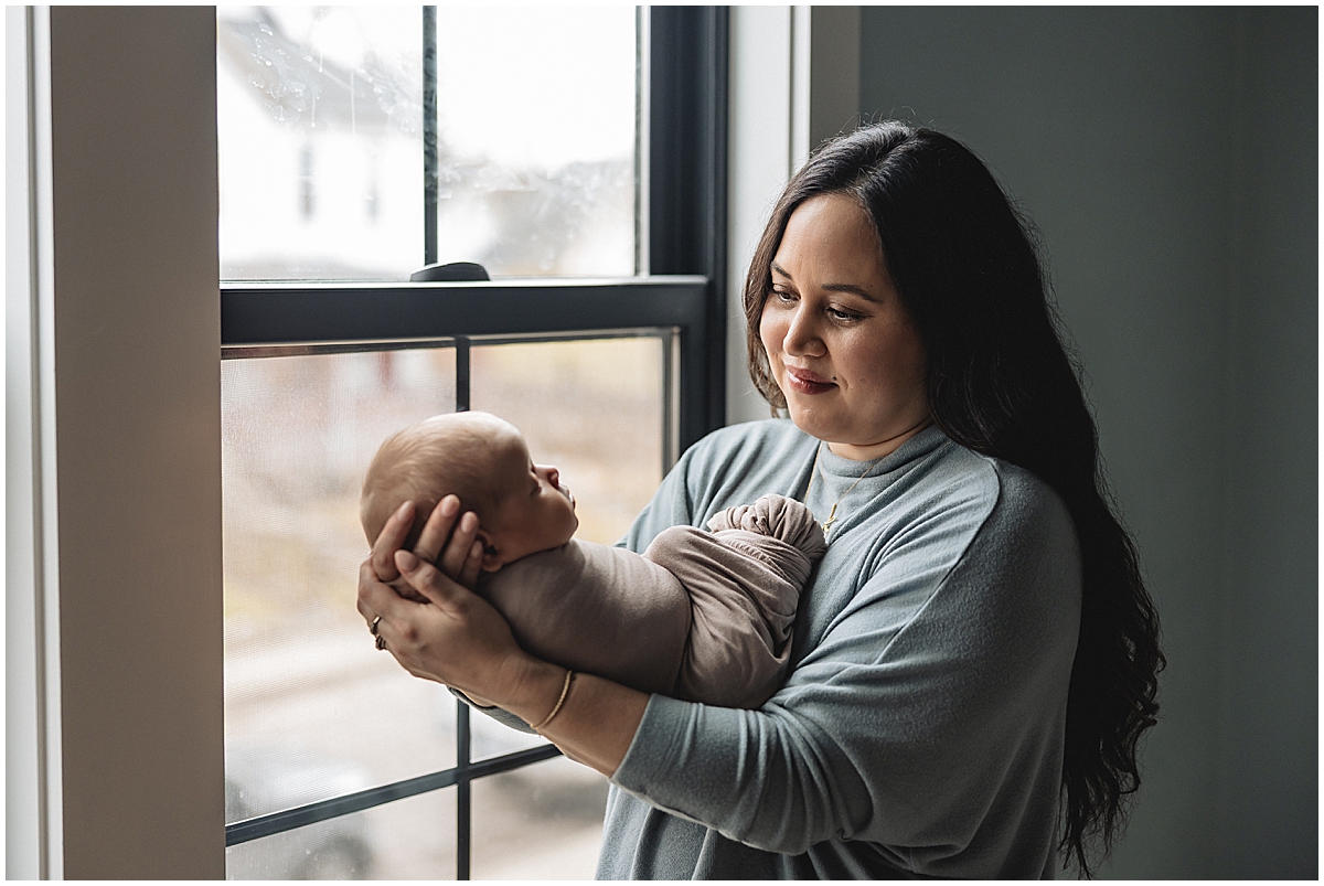 Mother in a soft blue outfit holding her swaddled newborn beside a window