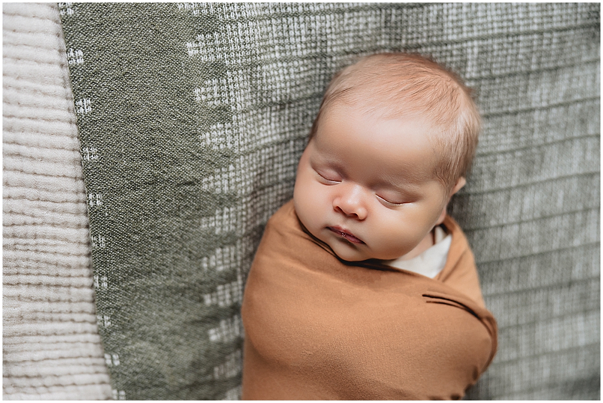 Sleeping newborn in a warm brown wrap against textured green and cream bedding