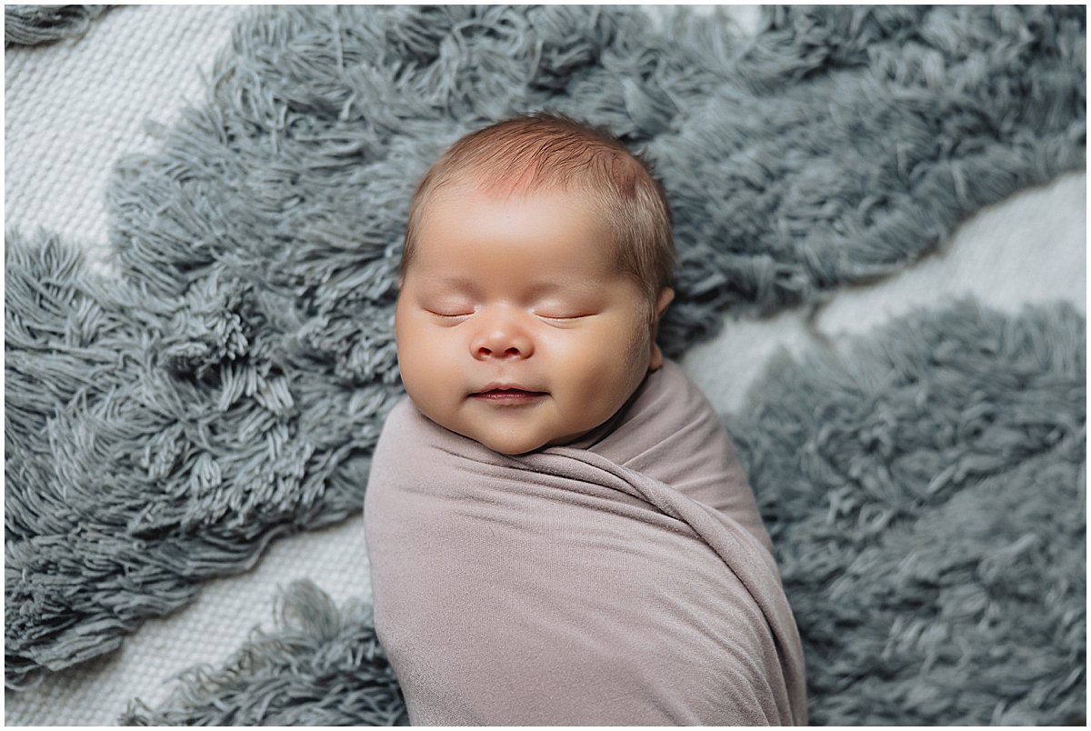 Sleeping newborn in a taupe wrap against a textured blue rug