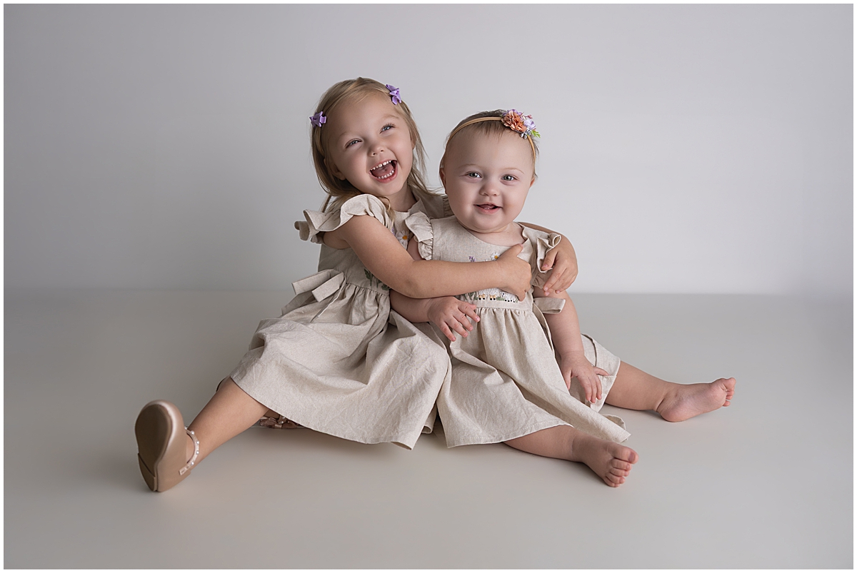 Big sister hugging the birthday girl as they sit together in matching lamb-embroidered dresses.